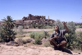 Image du Maroc Professionnelle de  Le paysage, formé par la kasbah Tifoultoute  qui se dresse majestueusement sur un éperon rocheux qui domine la vallée de l'oued Ouarzazate, attire les touristes qui prennent le temps de s'arrêter pour prendre une photo. Précisément à cet endroit un charmeur de serpent tenant une vipère avec sa main gauche tout en surveillant le mouvement du scorpion noir qu’il avait accroché à son cou avec sa main droite pose aux touristes contre un billet avec derrière lui comme décor celui qui avait servit pour de grand films comme Lawrence d’Arabie et Jésus de Nazareth à quelque kilomètres de la ville de Ouarzazate au sud du Maroc le Samedi 23 Août 1997. (Photo / Abdeljalil Bounhar) 